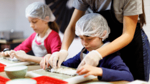 Two toddlers dressed in aprons and hats, learn to make sushi. The instructor is standing behind one of the kids and helping put the sushi rice on the seaweed sheets. This depicts a real life toddler cooking class, where toddlers are cooking with adult supervision and following a recipe. 