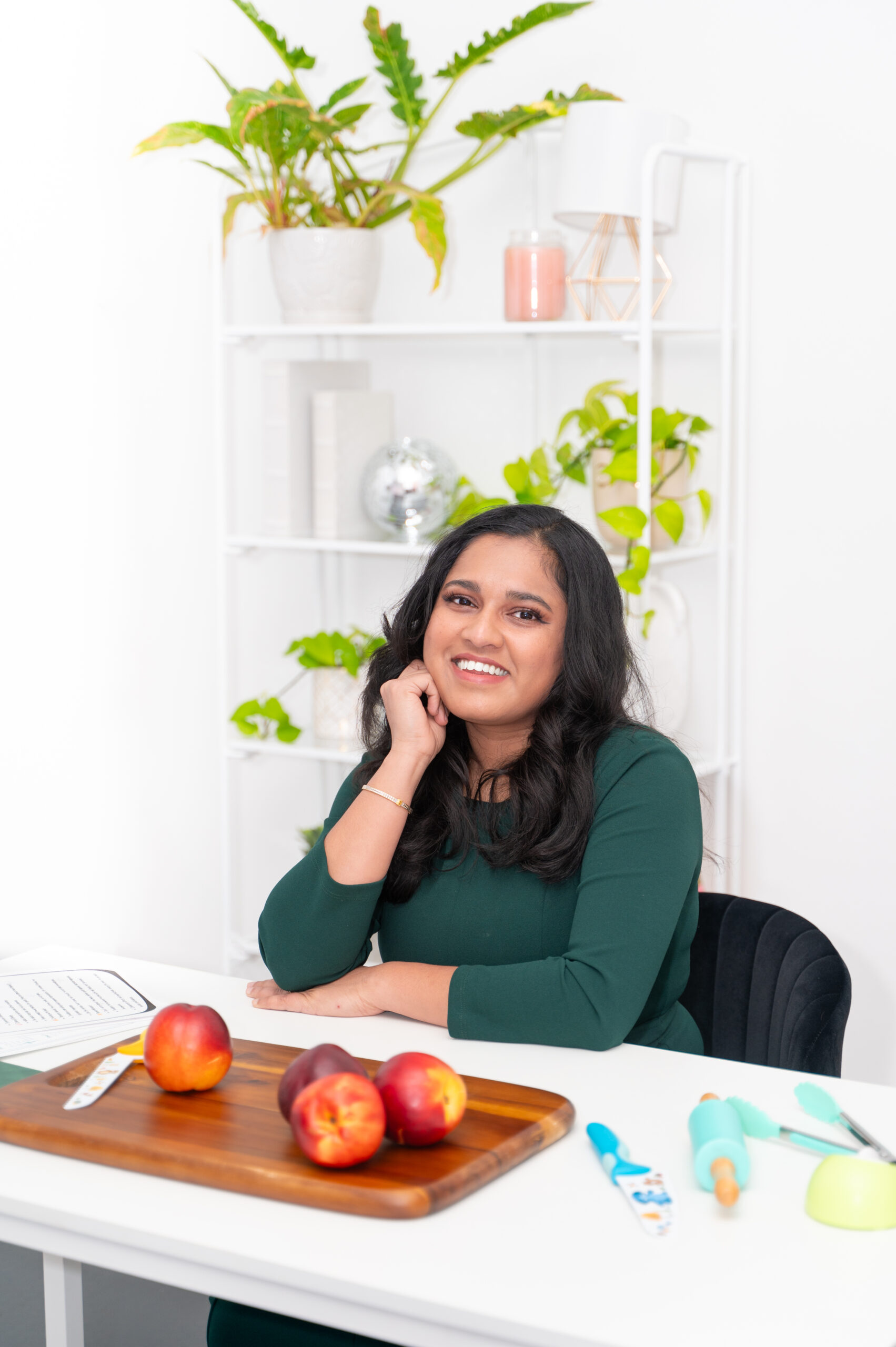 LAP_7767 A smiling woman in a green dress sits at a table with apples and baking tools. A white shelf with green plants and decor is in the background.