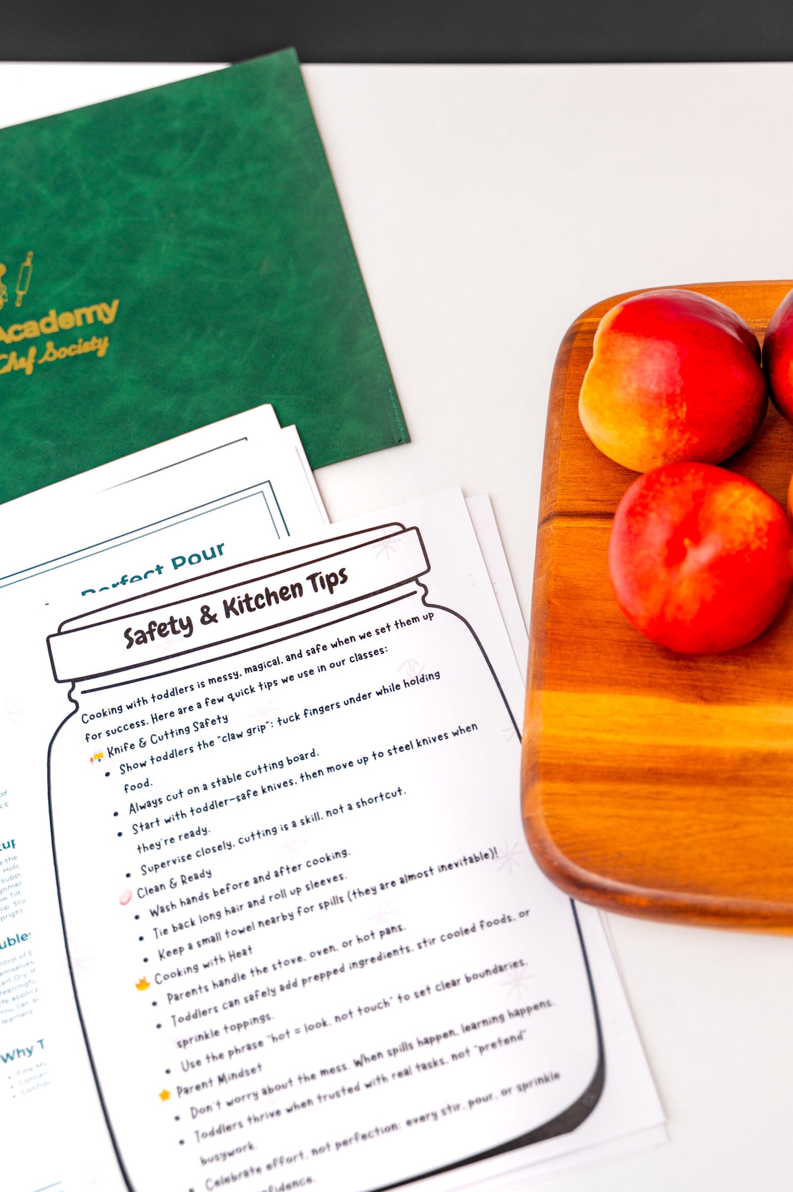 Cooking safety tips on a paper beside a green folder and three peaches on a wooden board. Bright, organized, and educational kitchen setting.
