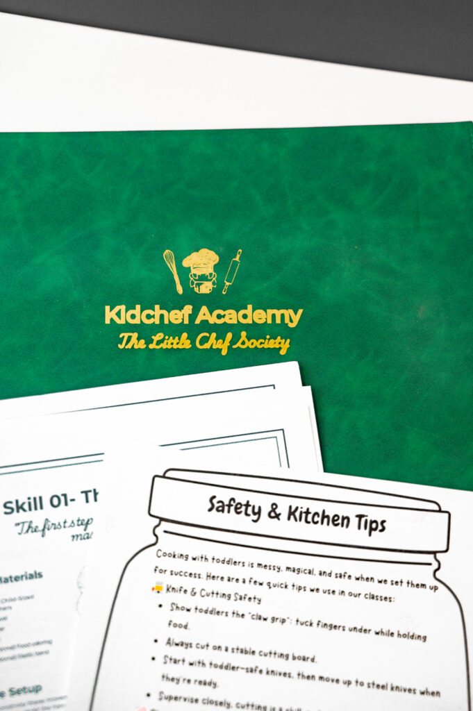 Home A green folder with "Kidchef Academy The Little Chef Society" in gold lettering. Below are papers titled "Safety & Kitchen Tips" and "Skill 01."